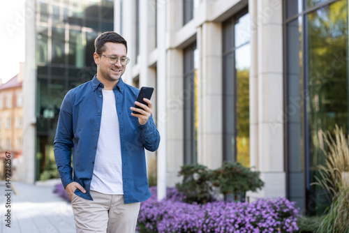 Fotografi A man in glasses using his phone while walking outside, enjoying a sunny day