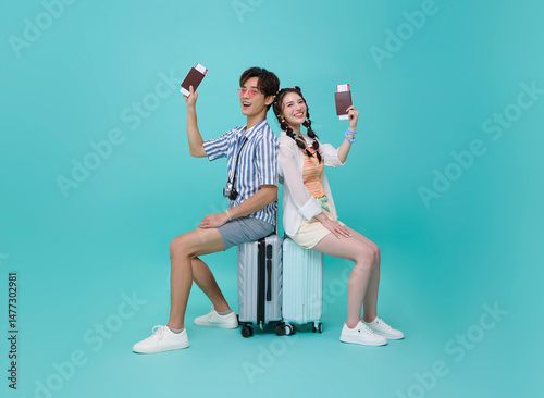 Happy smiling young Asian couple tourist sitting on luggage back to back and showing passport tickets going to travel on holidays isolated on blue background.