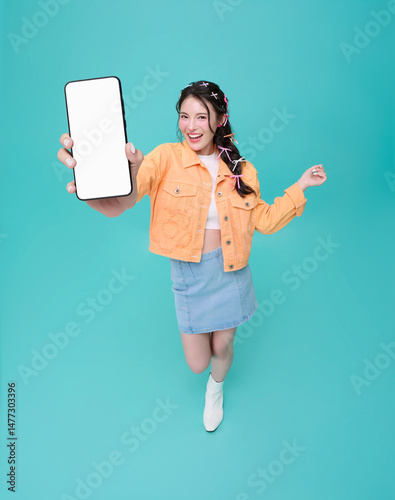 Happy beautiful young Asian teen woman showing smartphone mockup blank screen isolated on blue background.