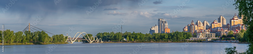 Obraz premium Modern residential buildings and pedestrian bridge on the Dnipro River in Kyiv at sunset. Peaceful cityscape with calm water, trees, and dramatic sky.