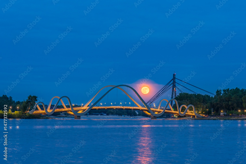 Obraz premium Full moon rises over the Dnipro River in Kyiv, glowing behind a modern pedestrian bridge. City lights and calm water create a peaceful and dreamy evening atmosphere.