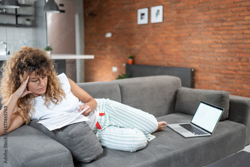 Obraz premium Stressed woman reading documents on sofa with laptop