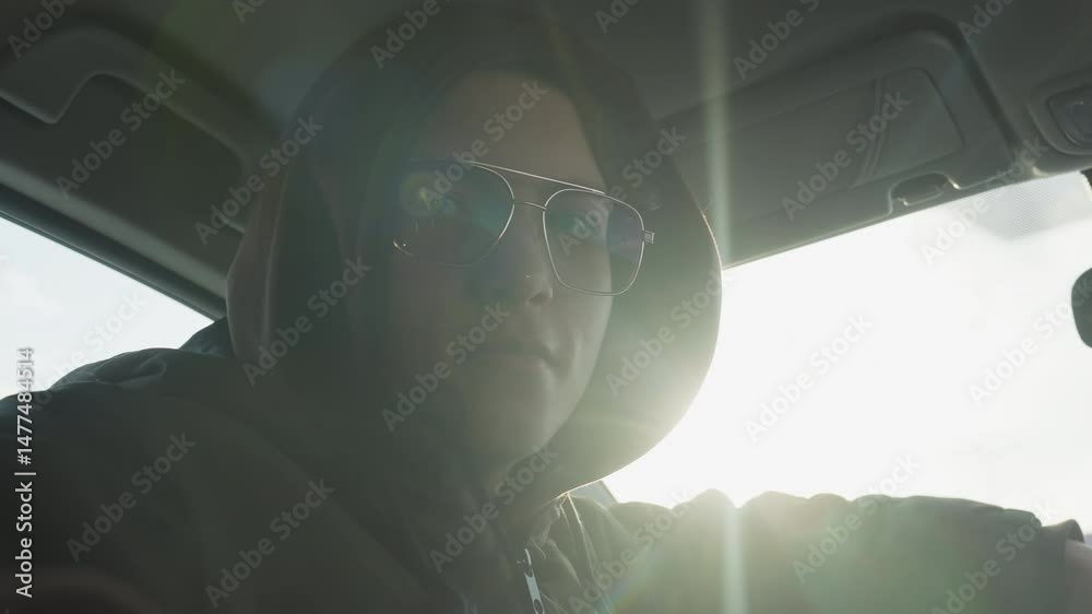 student wearing hoodie and tinted glasses turns head back confidently over shoulder while gripping steering wheel inside sunlit car cabin overlooking snowy urban background