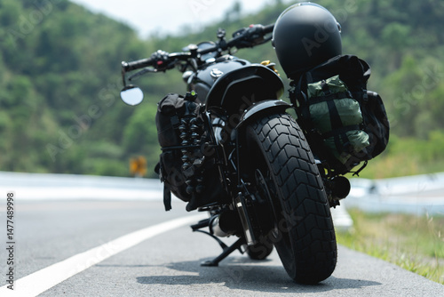 A black motorcycle loaded with gear and a helmet rests on a winding road with lush green hills in the background.