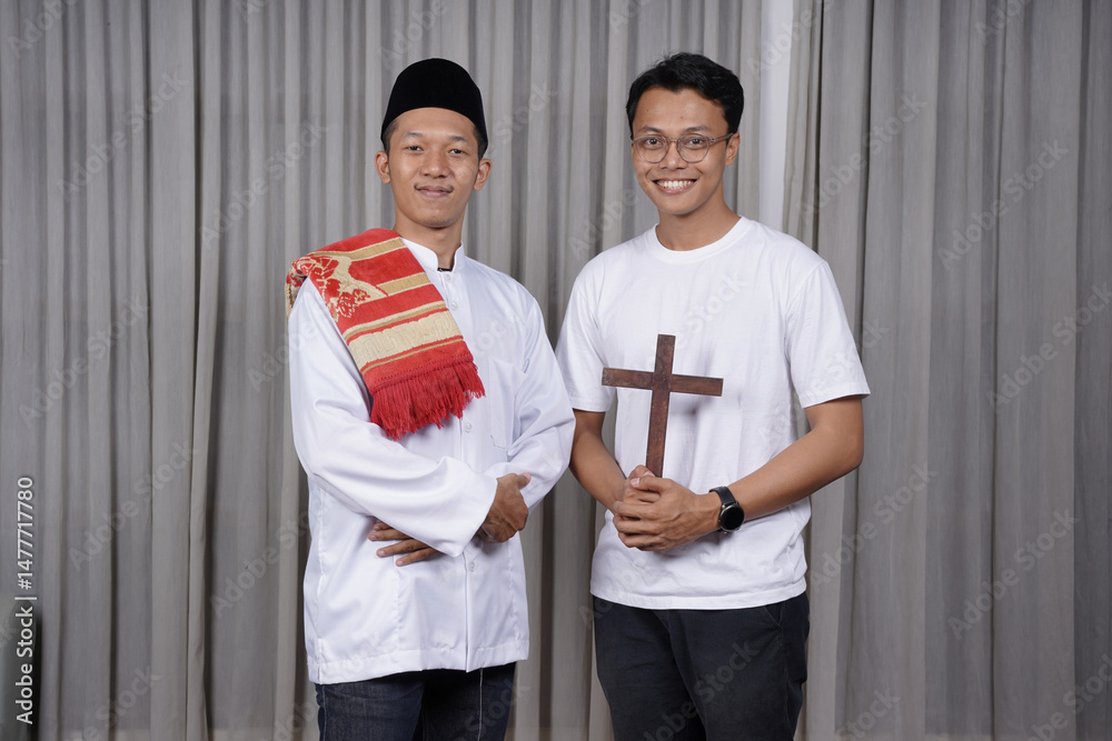 Φωτογραφία A Muslim man wearing a white koko shirt, a peci and carrying a prayer mat on his shoulder stands side by side with a Christian man carrying a cross in his hand