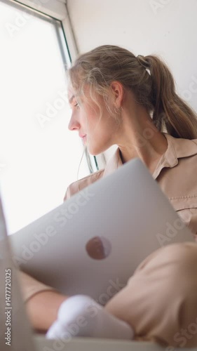 Young copywriter woman looks through window to distract from working on texts on laptop behind brown curtain in comfortable room slow motion