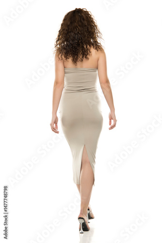 Back view of woman with curly brown hair in beige strapless dress with back slit walking in clear heels against white background, elegant fashion portrait