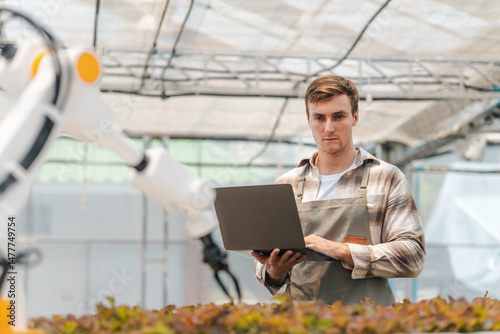 Male farmer using laptop in greenhouse to check the operation of robotic arm with AI system to control in farm care. Smart agriculture technology concept. Wireless robot for agriculture in smart farm.