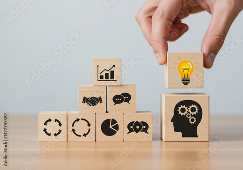 A hand adds a light bulb icon cube (representing ideas) to a stack of business concept cubes, next to a head with gears (representing thinking).
