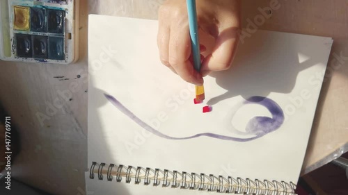 Artist's hand painting abstract design with watercolors in sunlit studio.