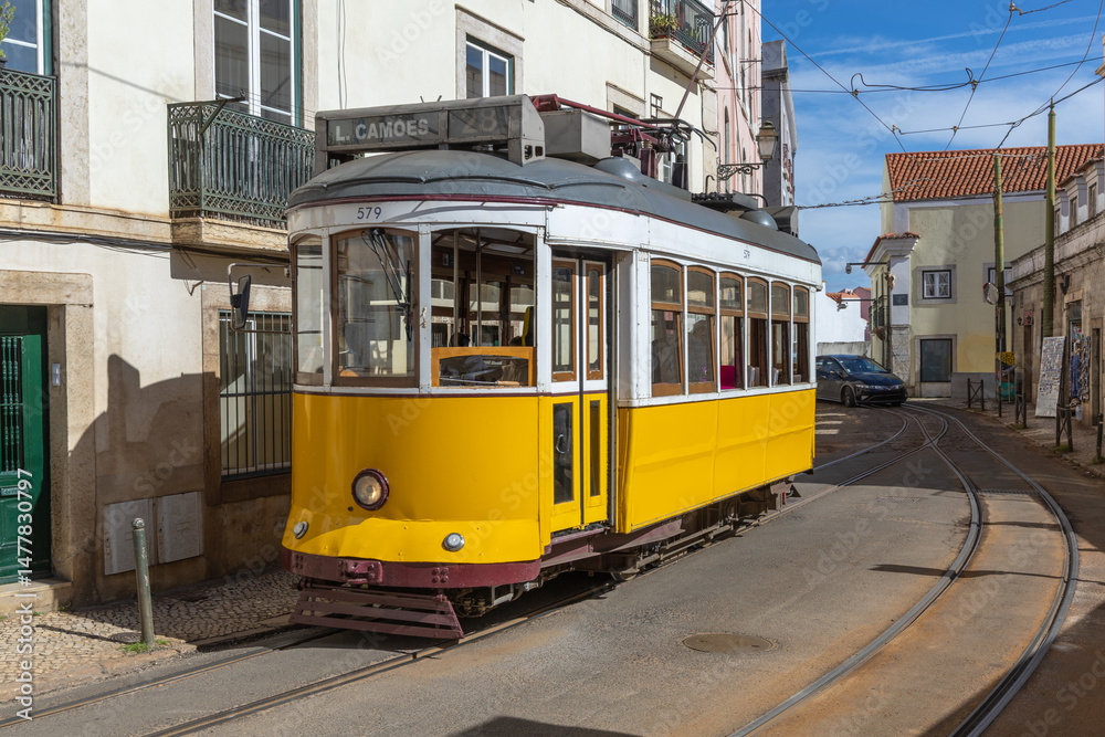 Naklejka premium A classic yellow Tram 28 navigates the charming, narrow streets of Lisbon, Portugal on a sunny day.