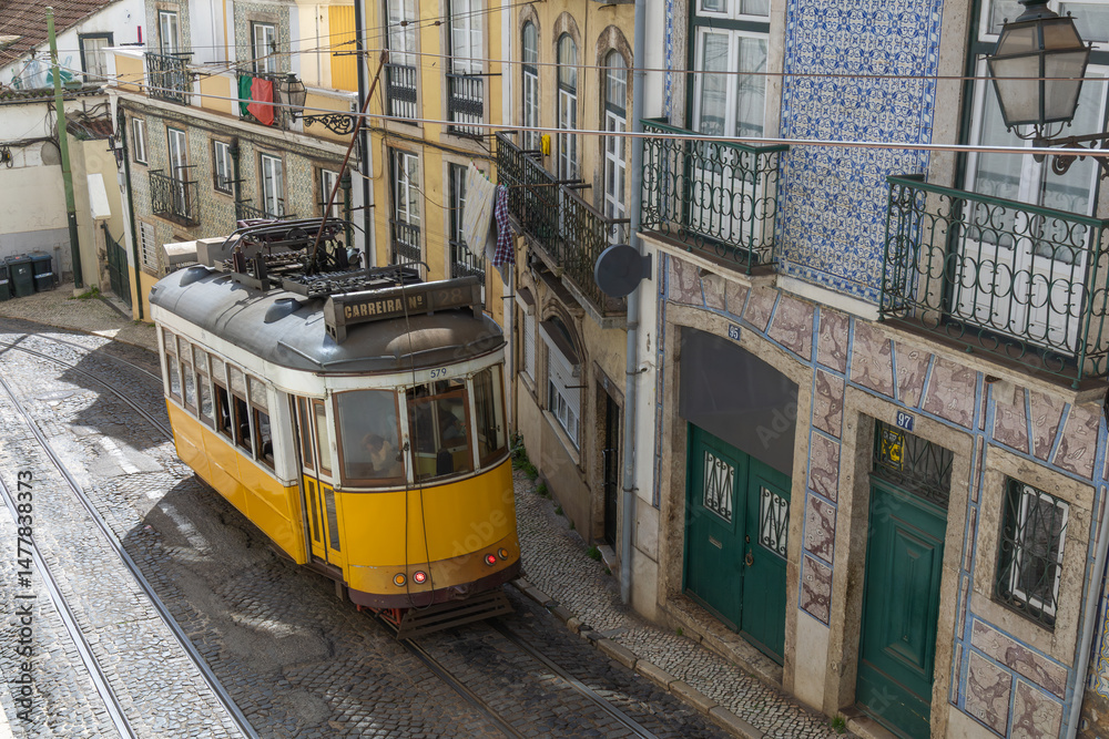 Φωτογραφία Historic yellow tram navigating a narrow, cobbled street in Lisbon, Portugal, with colorful tiled buildings and balconies in the background