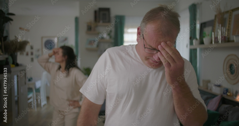 Obraz premium Man covering face with hands in frustration during argument, woman in background in living room, dramatic relationship conflict, focus on emotional struggle