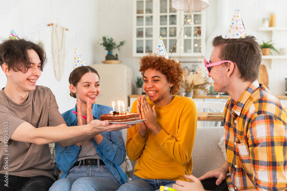 Obraz premium Make a wish. Woman wearing party cap blowing out burning candles on birthday cake. Happy Birthday party. Group of friends wishes girl happy birthday. People celebrating birthday with party at home