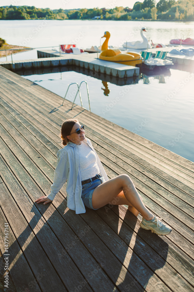 Beautiful blonde woman on the pier, wearing a white shirt, short denim shorts and sunglasses, cheerful woman on the river bank, summer beach vacation on the water.