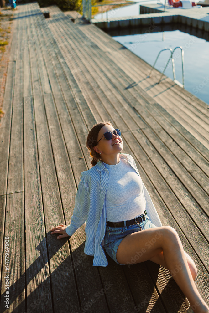 Beautiful blonde woman on the pier, wearing a white shirt, short denim shorts and sunglasses, cheerful woman on the river bank, summer beach vacation on the water.