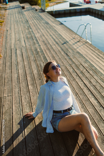 Obraz na płótnie Beautiful blonde woman on the pier, wearing a white shirt, short denim shorts and sunglasses, cheerful woman on the river bank, summer beach vacation on the water. w salonie
