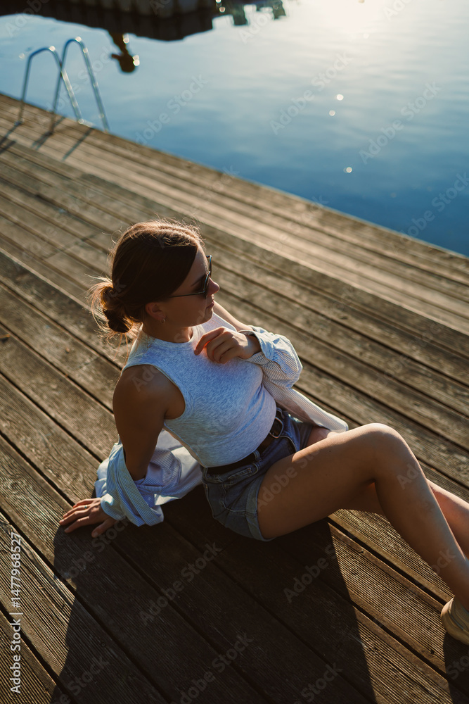 Beautiful blonde woman on the pier, wearing a white shirt, short denim shorts and sunglasses, cheerful woman on the river bank, summer beach vacation on the water.