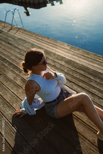 Beautiful blonde woman on the pier, wearing a white shirt, short denim shorts and sunglasses, cheerful woman on the river bank, summer beach vacation on the water.