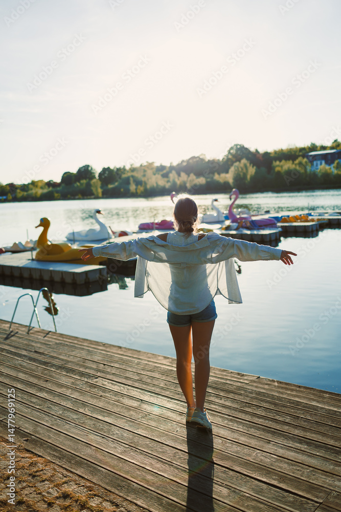 Beautiful blonde woman on the pier, wearing a white shirt, short denim shorts and sunglasses, cheerful woman on the river bank, summer beach vacation on the water.