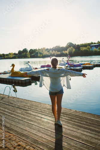 Obraz na płótnie Beautiful blonde woman on the pier, wearing a white shirt, short denim shorts and sunglasses, cheerful woman on the river bank, summer beach vacation on the water. w salonie