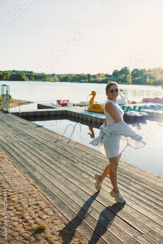 Obraz na płótnie Cheerful woman in shorts and sunglasses walking along the pier near the lake. Woman having fun and running under the scorching sun. Young woman on a walk near the lake. w salonie