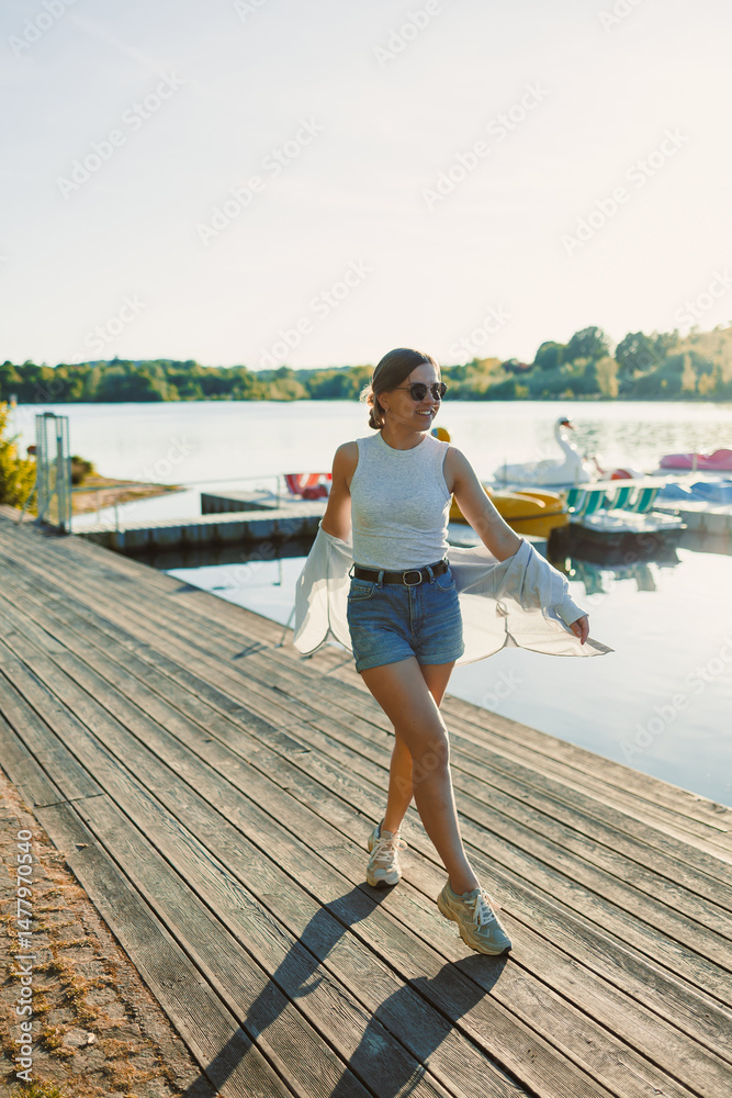 Cheerful woman in shorts and sunglasses walking along the pier near the lake. Woman having fun and running under the scorching sun. Young woman on a walk near the lake.