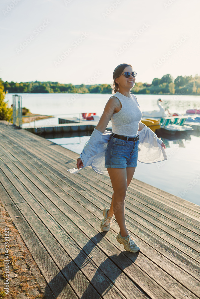 Cheerful woman in shorts and sunglasses walking along the pier near the lake. Woman having fun and running under the scorching sun. Young woman on a walk near the lake.