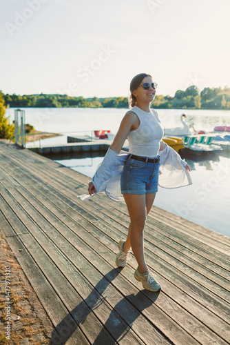 Obraz na płótnie Cheerful woman in shorts and sunglasses walking along the pier near the lake. Woman having fun and running under the scorching sun. Young woman on a walk near the lake. w salonie