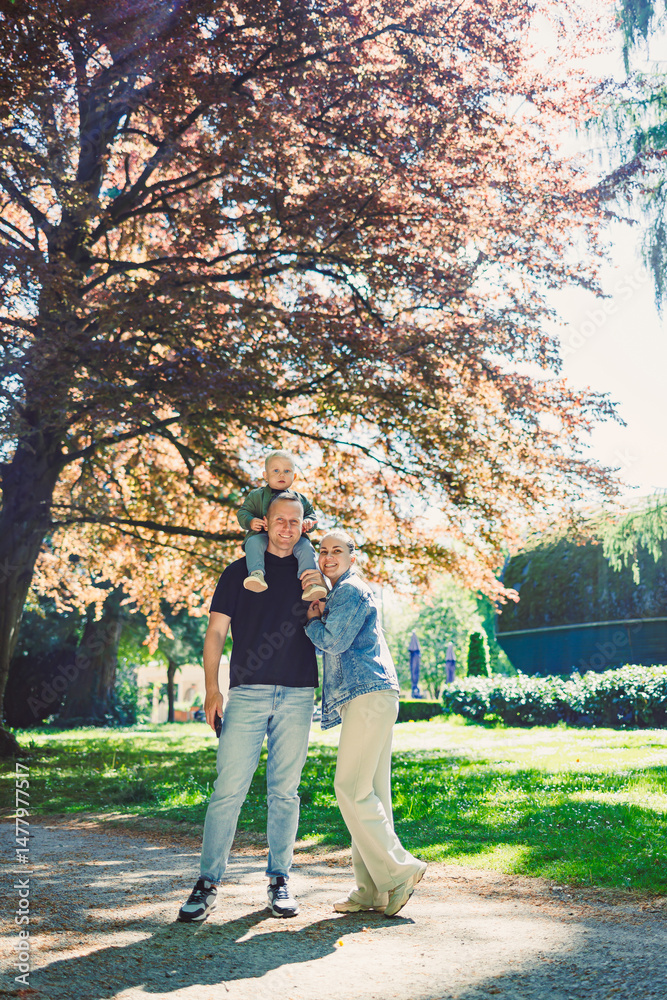 A Truly Joyful Family Moment Captured Underneath a Beautiful Tree in a Sunlit Park Where a Little Child Is Happily Enjoying Being on Their Parents Shoulders, Filling Hearts with Love and Happiness