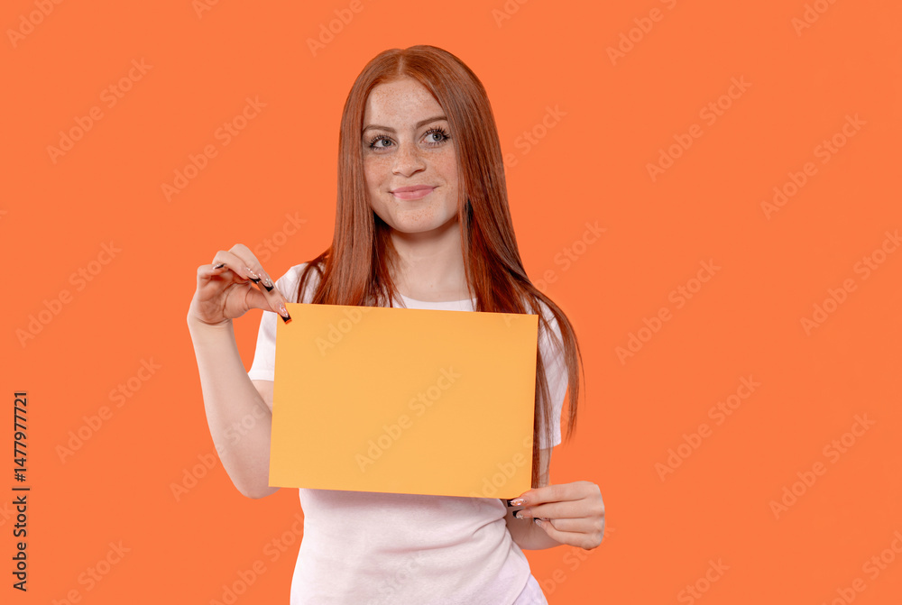 Young woman with long red hair holds blank orange paper in front of solid orange background during a creative studio session in the afternoon Young woman with long red hair holds blank orange paper in front of solid orange background during a creative studio session in the afternoon