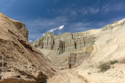 Stunning desert landscape featuring multicolored, striated hills and eroded rock formations beneath a dramatic sky with heavy, moody clouds. The stark contrast between the bright yellowish-beige deser