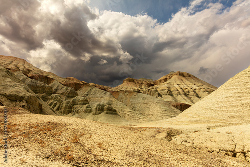 Stunning desert landscape featuring multicolored, striated hills and eroded rock formations beneath a dramatic sky with heavy, moody clouds. The stark contrast between the bright yellowish-beige deser