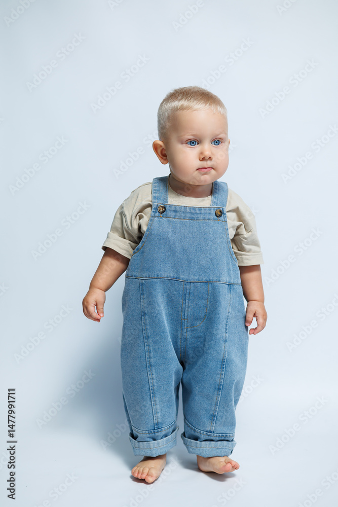 Adorable and cute baby dressed in denim overalls is posing adorably against a light and soft background, showcasing a stylish look that highlights the charm of childhood and innocence
