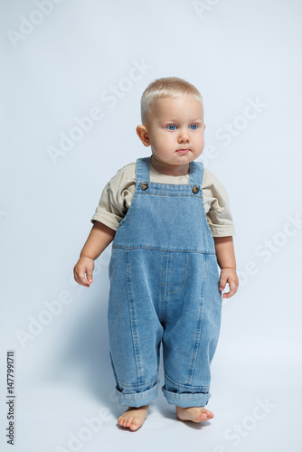 Obraz na płótnie Adorable and cute baby dressed in denim overalls is posing adorably against a light and soft background, showcasing a stylish look that highlights the charm of childhood and innocence w salonie