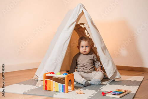 An adorable little toddler is joyfully playing inside a cozy tent, surrounded by a variety of colorful toys, all set in a bright and cheerful indoor environment that radiates warmth and fun