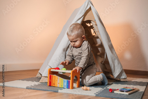 Obraz na płótnie An absolutely adorable baby is joyfully playing with a colorful musical toy while nestled inside a cozy indoor tent, creating a warm and inviting atmosphere for exploration and fun w salonie