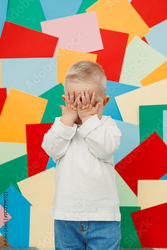 Obraz na płótnie A delightful child is playing, hiding their face behind their hands in a cheerful and playful expression, set against a vibrant and colorful background filled with various shapes and patterns w salonie