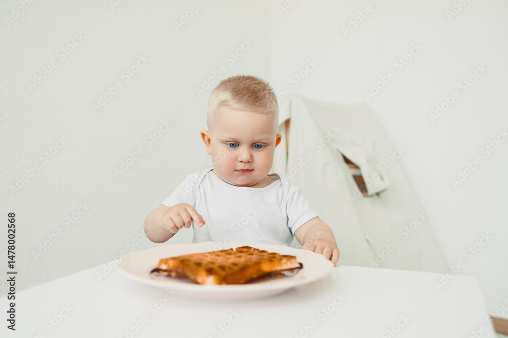 An adorable baby, full of curiosity, is examining a delicious waffle placed on a bright white table within a bright, minimalistic setting, creating a delightful scene of childhood exploration