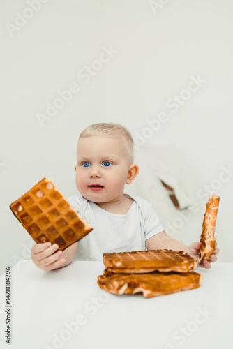 Obraz na płótnie An adorable baby is joyfully exploring and playing with small pieces of waffles spread out on a clean white table, showcasing a delightful moment of curiosity and creativity during mealtime w salonie