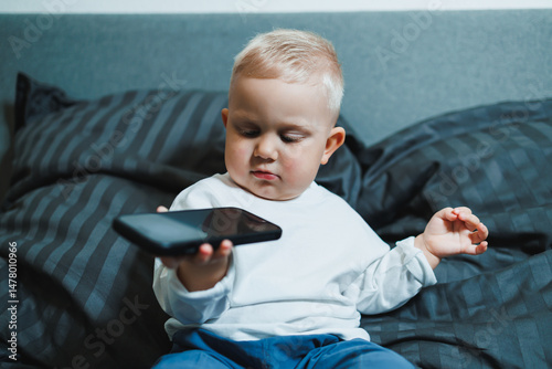 Obraz na płótnie A curious toddler is happily engaged with a smartphone while comfortably nestled in a cozy bed surrounded by soft cushions, embodying the innocence and carefree spirit of childhood and playtime w salonie