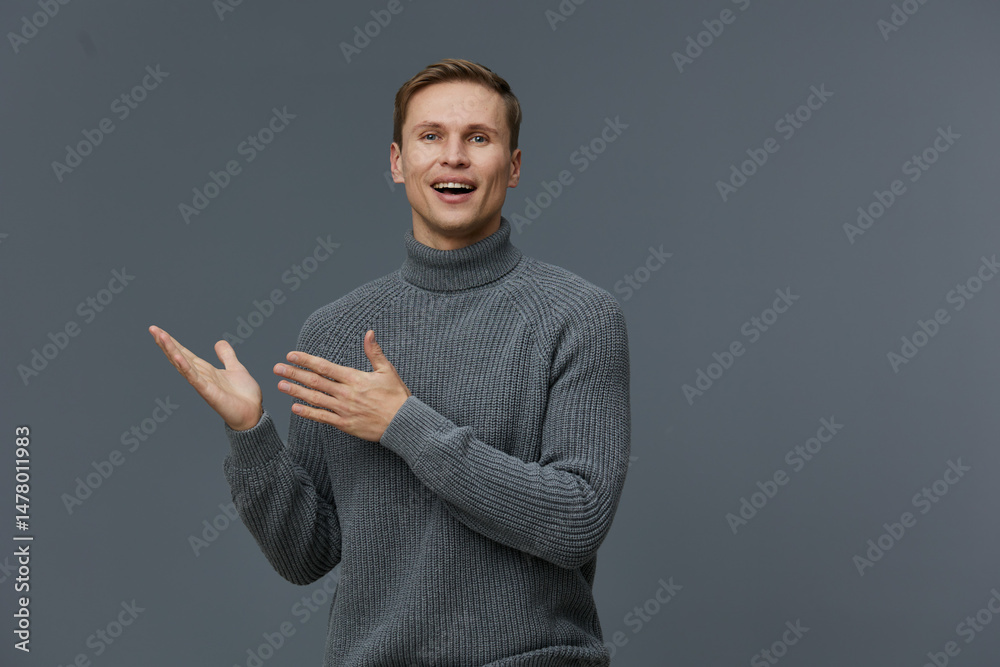 Fototapeta premium Confident young man in cozy gray sweater gesturing with hands and smiling cheerful at camera on plain gray background, studio portrait. People lifestyle concept