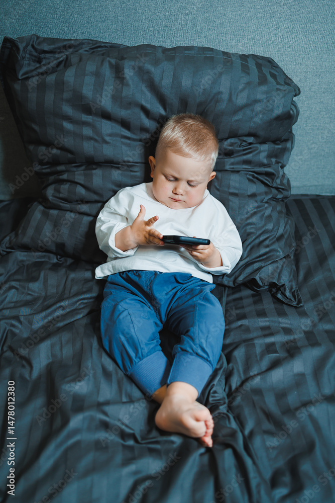 A young toddler is comfortably engaged with a smartphone while sitting on a cozy bed, demonstrating a sense of curiosity and focus during their leisure time spent indoors in a family environment