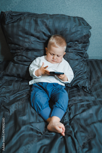 Obraz na płótnie A young toddler is comfortably engaged with a smartphone while sitting on a cozy bed, demonstrating a sense of curiosity and focus during their leisure time spent indoors in a family environment w salonie