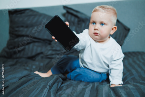 A curious toddler is sitting comfortably on a bed in a cozy bedroom environment while holding a smartphone, exploring the digital world with a sense of wonder and engaging in playful interaction