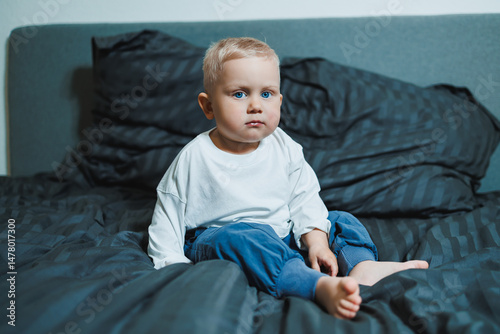 Obraz na płótnie An adorable toddler is sitting on a bed, showcasing a curious expression that captures the enchanting essence of childhood in a cozy bedroom setting filled with warmth and comfort w salonie