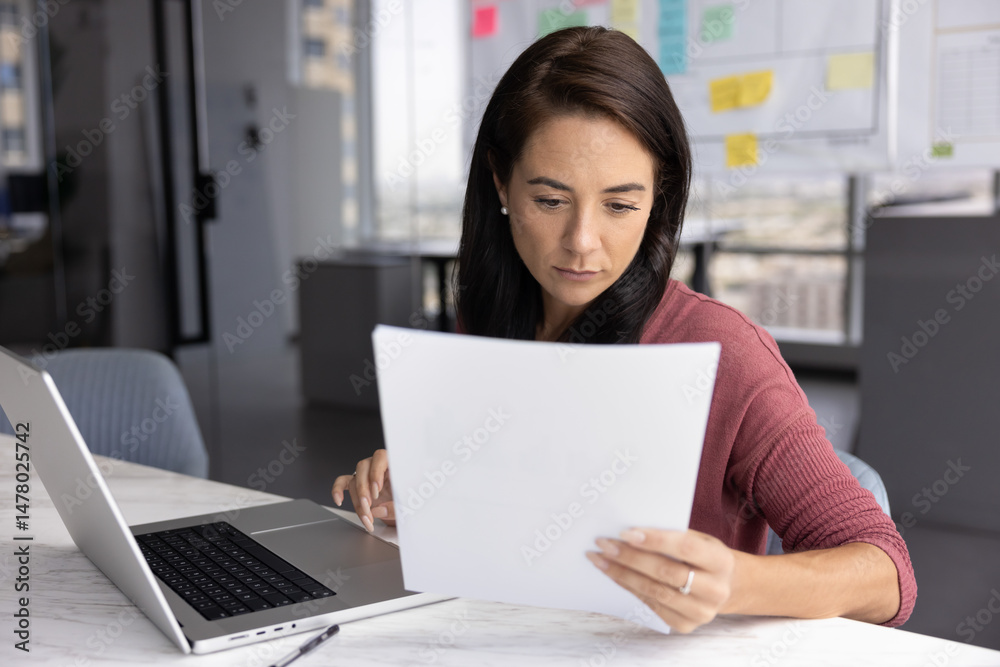 Paperwork for productivity. Serious young businesswoman project manager accountant prepare financial or statistical report at office desk review paper documents to fill in electronic form on notebook Paperwork for productivity. Serious young businesswoman project manager accountant prepare financial or statistical report at office desk review paper documents to fill in electronic form on notebook