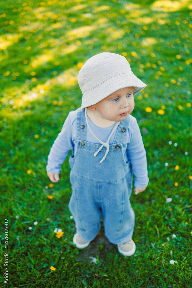 An adorable child dressed in denim overalls and wearing a cute sun hat is joyfully playing on a bright, sunny day in a vibrant green field filled with fluffy dandelions swaying gently in the breeze