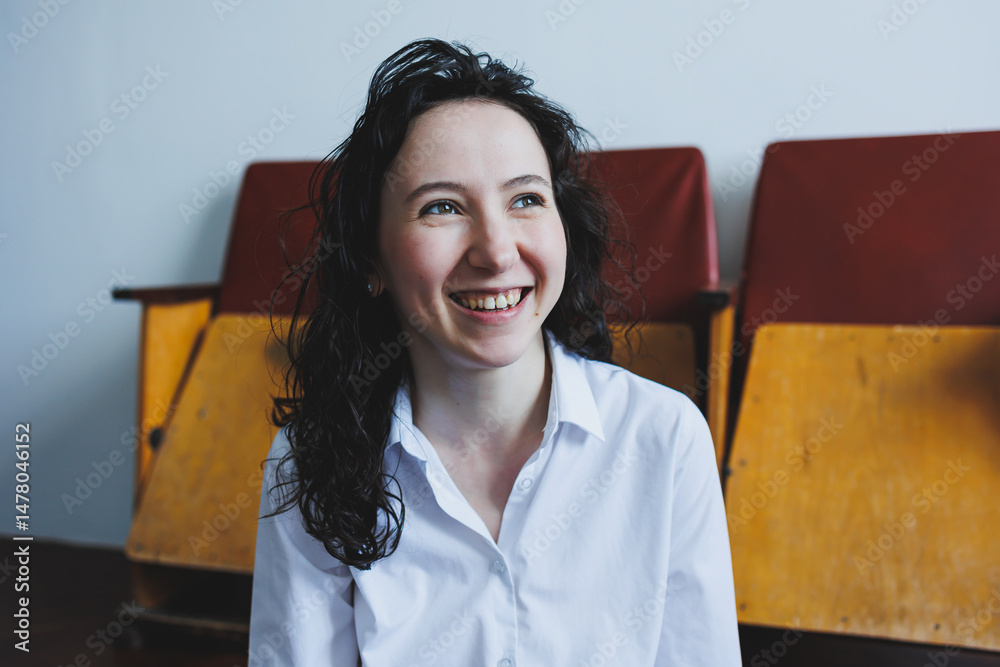 A cheerful young woman is seen smiling brightly in a minimalist indoor setting adorned with vintage chairs, creating a warm and inviting atmosphere that radiates joy and confidence