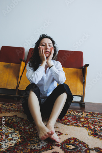 A contemplative woman dressed in casual attire is seated on a vibrant and colorful rug situated within the confines of a minimalist room, exuding an air of tranquility and peacefulness